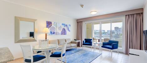 A modern living room with a glass dining table, white chairs, a sofa, and a blue armchair. There is a large window with curtains, a flat-screen TV, and artwork on the wall.