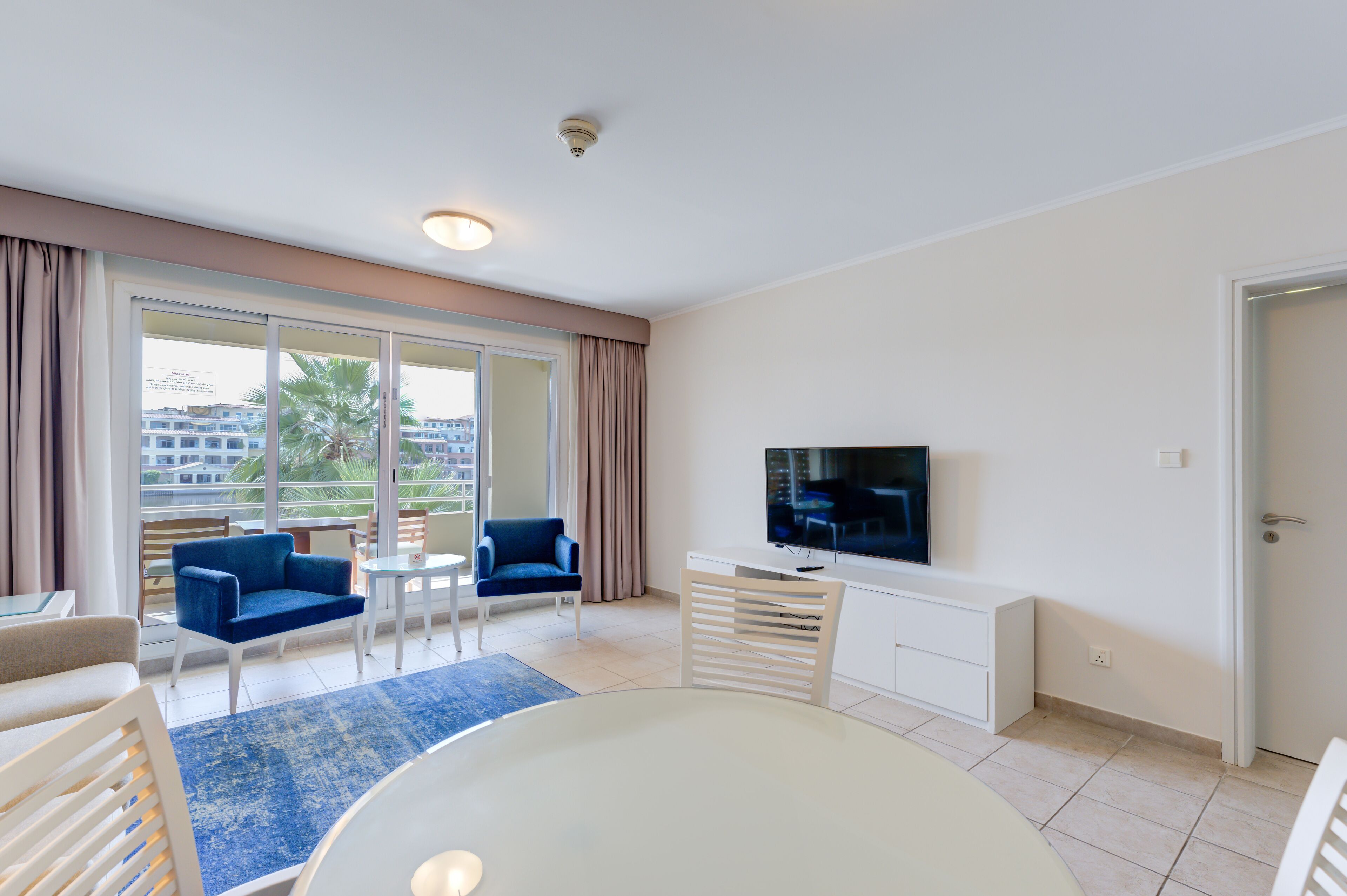 A modern hotel room with a large window, a flat-screen TV, a white desk, and a blue sofa.