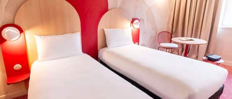 Habitación de hotel con dos camas, una pared de acento roja, una mesita con una silla y una ventana con cortinas.