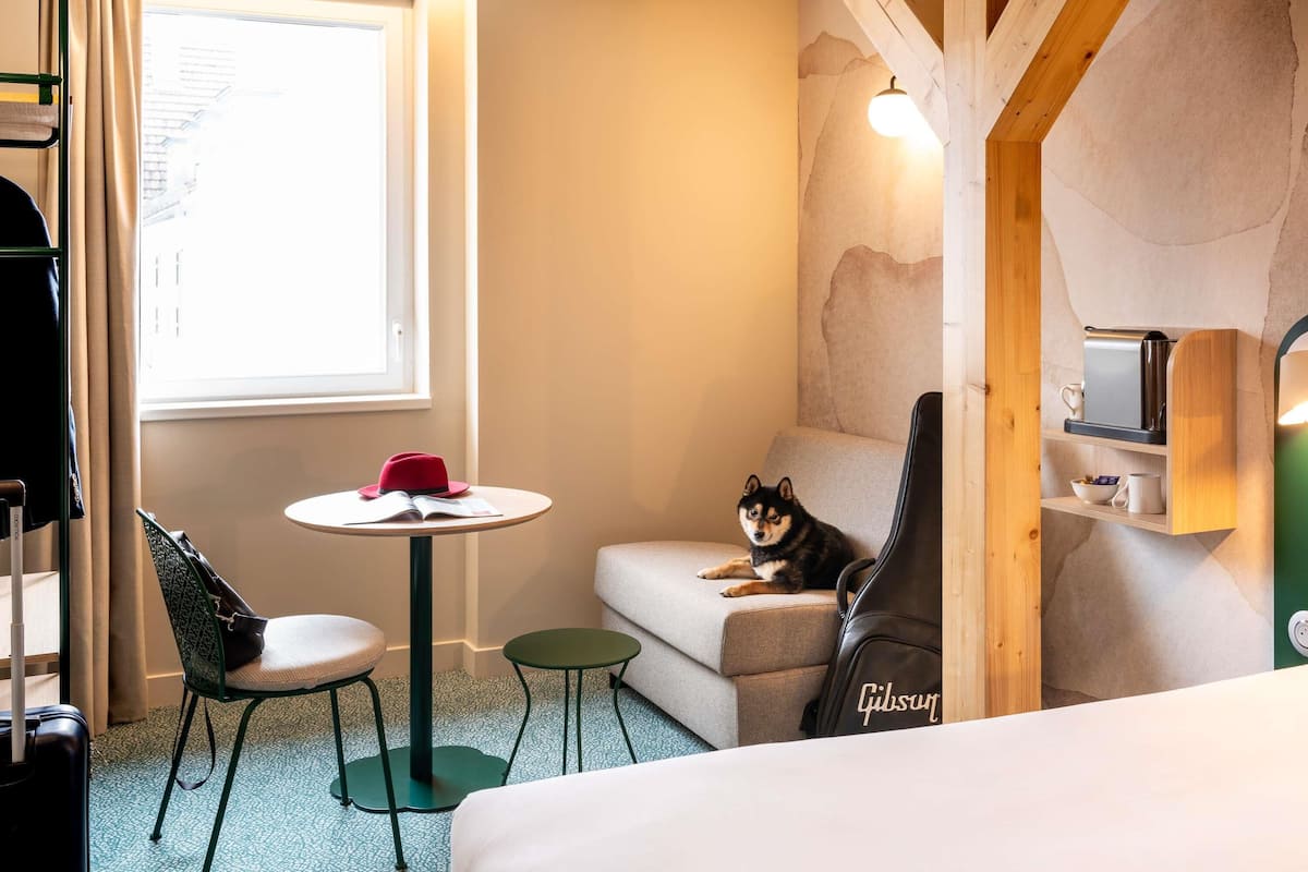 A hotel room with a bed, a small round table with a red hat, a green chair, a sofa, and a guitar.