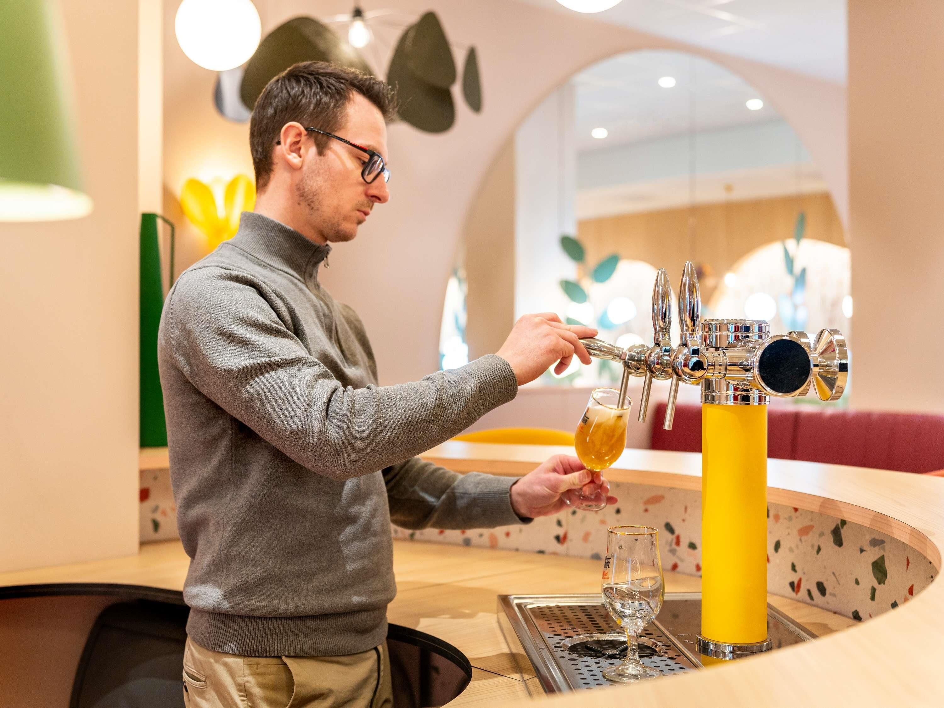 Una persona servint cervesa a un got a la barra d'un bar amb un disseny modern, que inclou un pilaret groc i una zona de seients amb un sofà vermell.