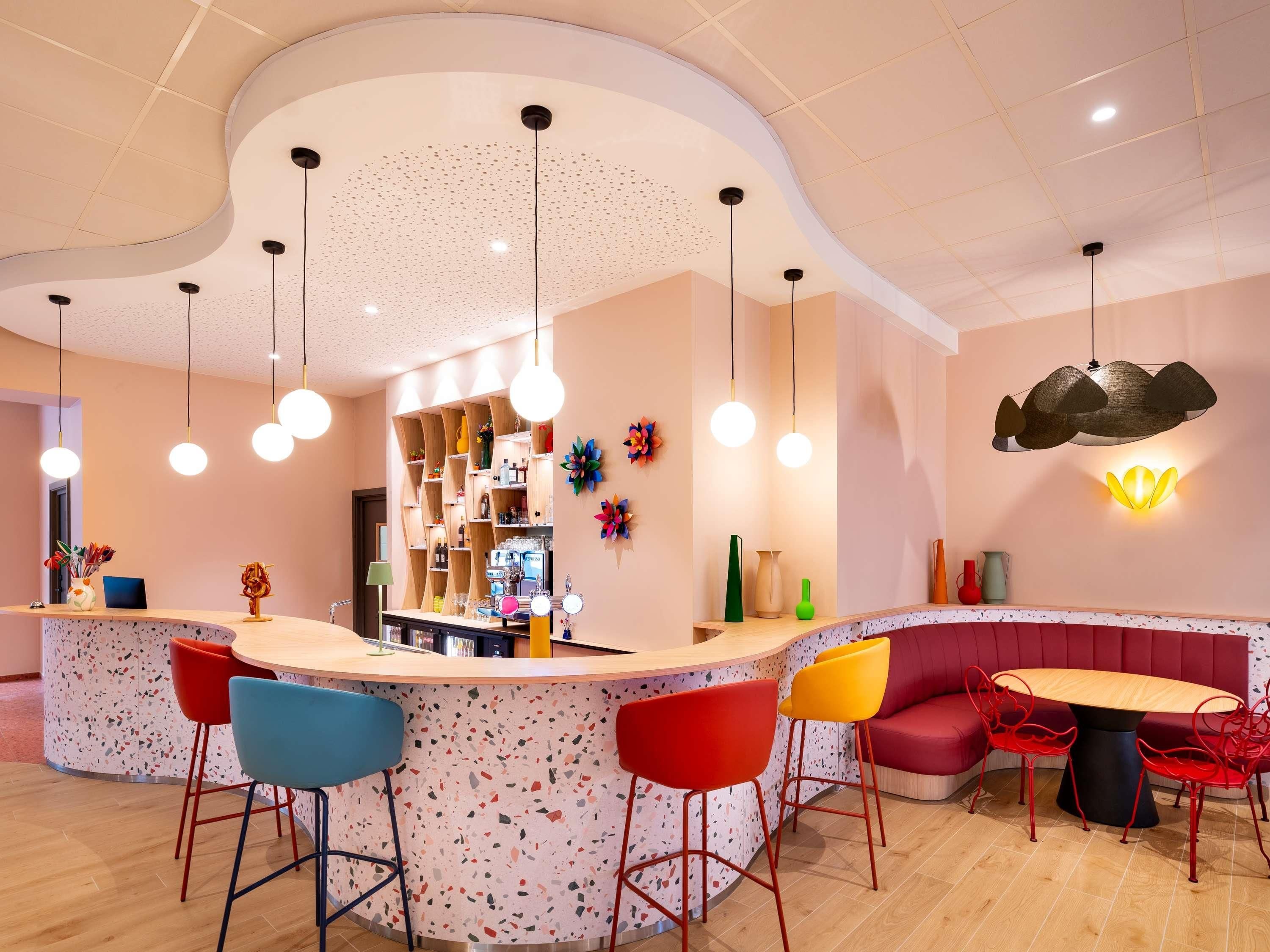 Een moderne bar met een gebogen toonbank, kleurrijke barkrukken en een zithoek met een ronde tafel en rode stoelen.