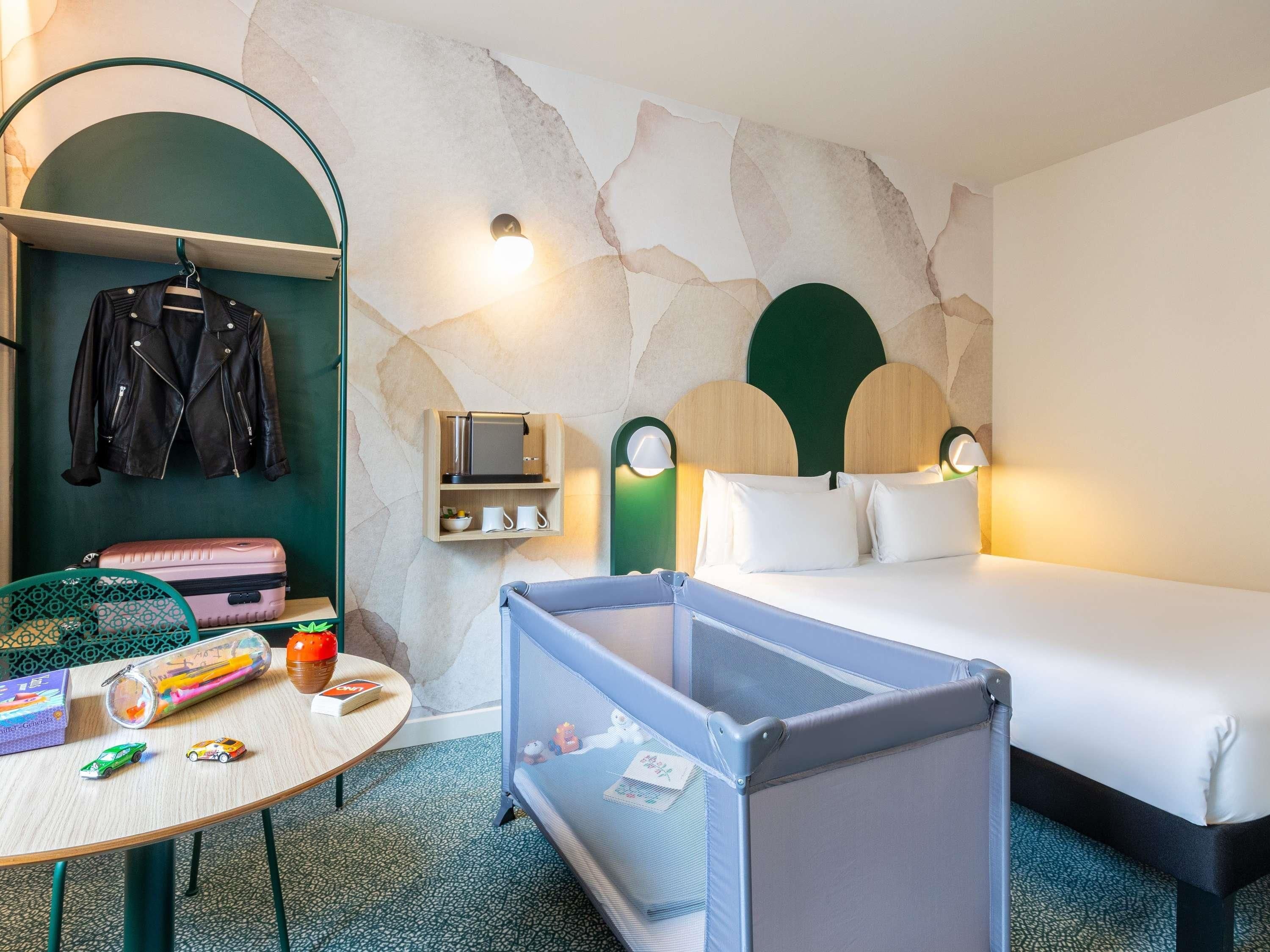 Quarto de hotel com uma cama, uma escrivaninha com cadeira, um cabideiro, uma pequena mesa com brinquedos e um berço para bebê.
