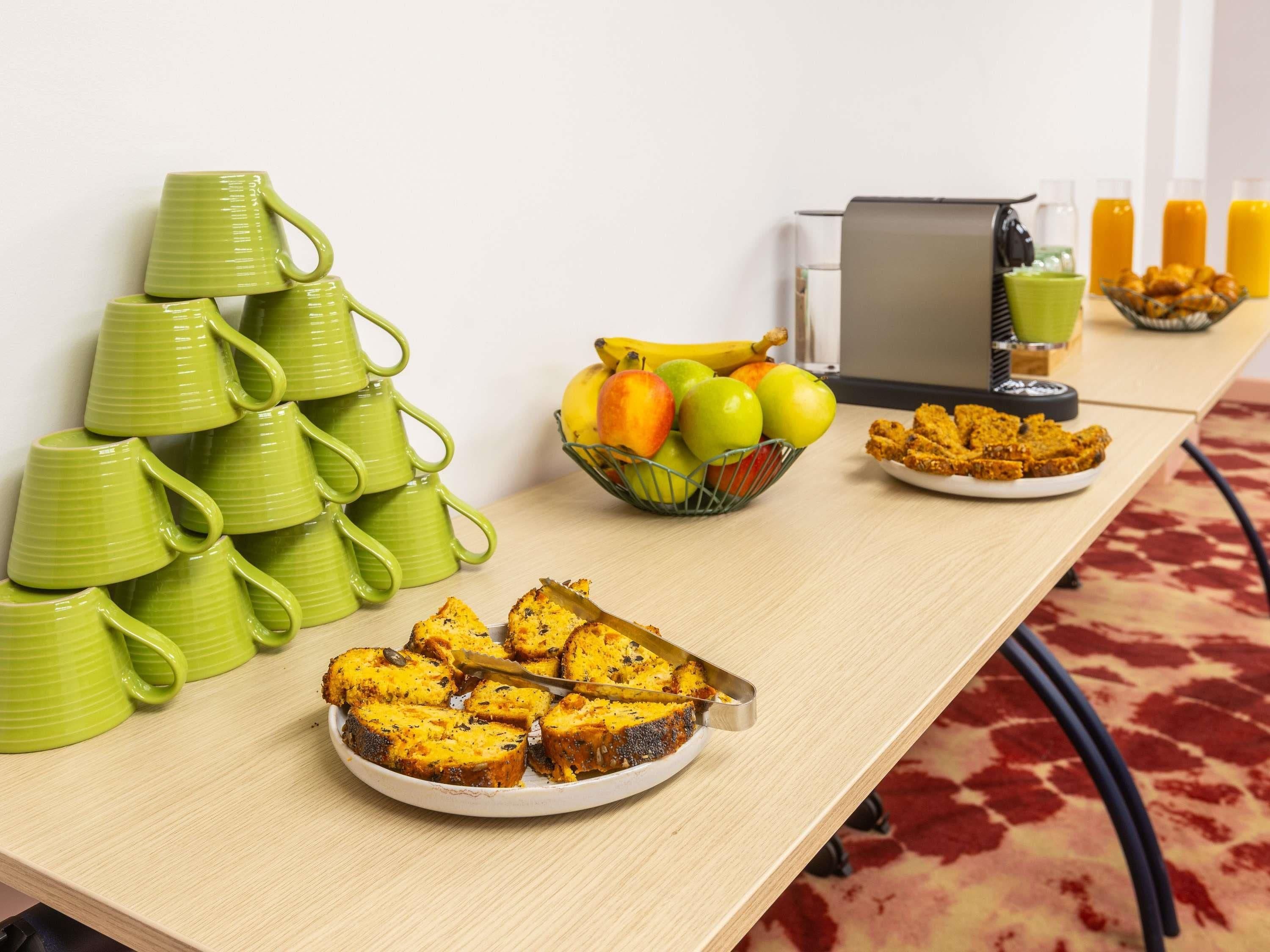 A breakfast buffet setup with green mugs, a fruit bowl, a plate of muffins, and juice bottles.