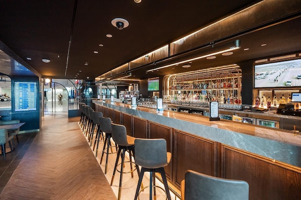 A modern bar with a long counter, bar stools, and a digital screen displaying a sports game.