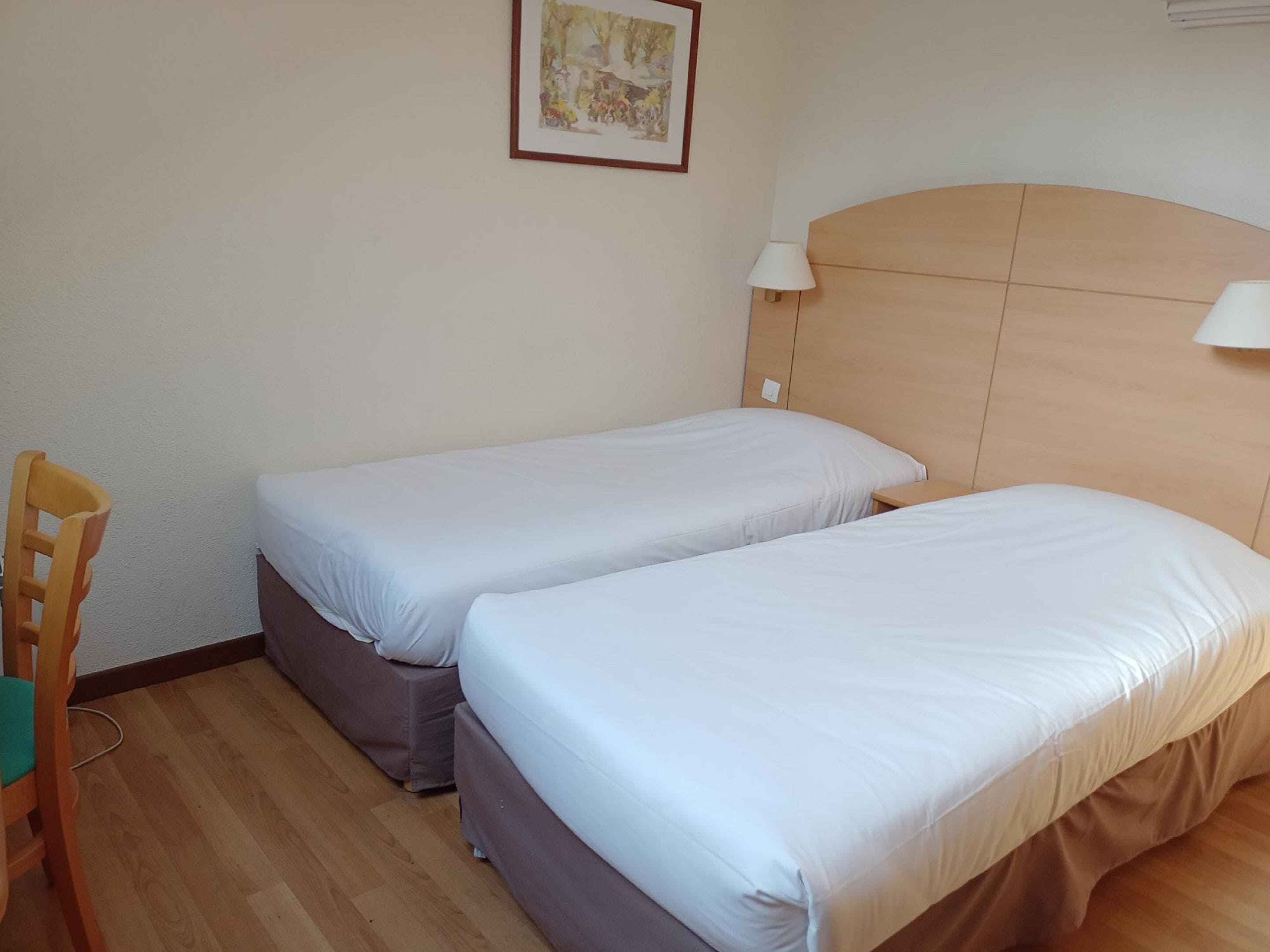 Una habitación con dos camas individuales, una silla de madera, una mesita pequeña y una lámpara de pared.