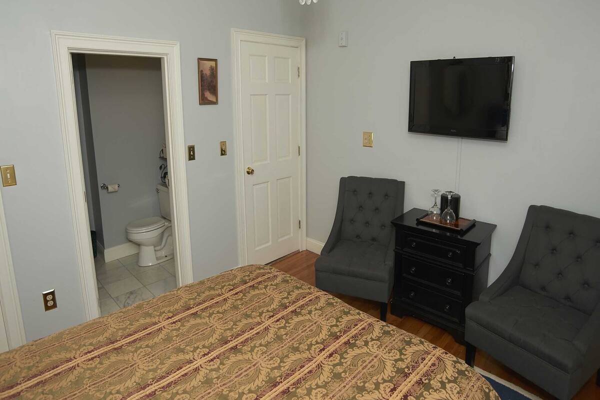 A bedroom with a bed, two armchairs, a TV, and a dresser.