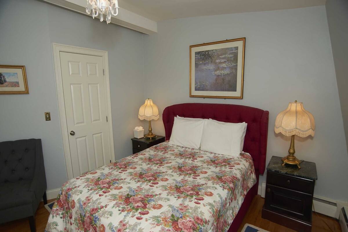 A bedroom with a floral bedspread, a red upholstered headboard, two bedside lamps, a framed painting on the wall, and a door with a small picture frame above it.