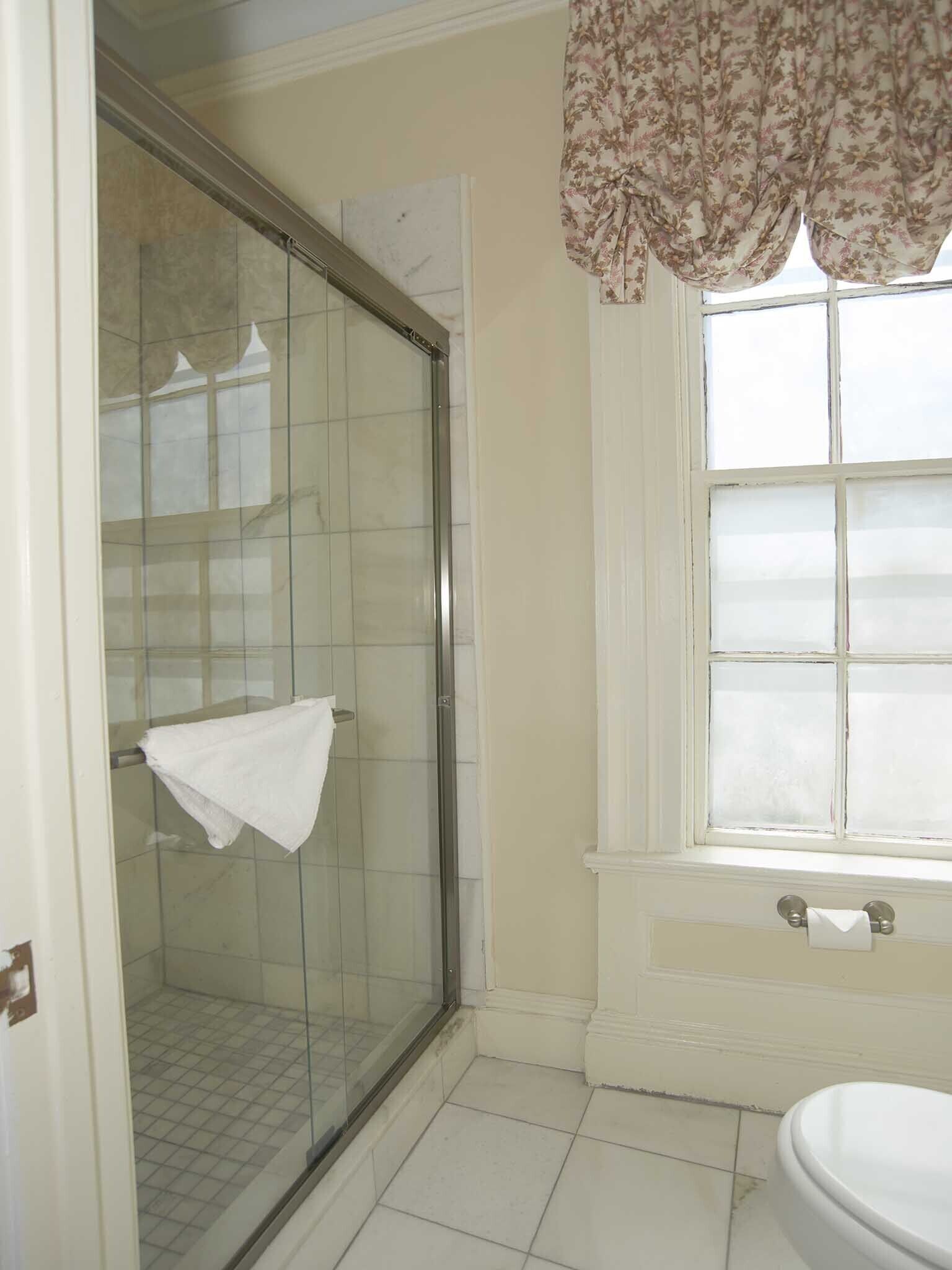 A bathroom with a glass shower enclosure, a toilet, and a window with a floral curtain.