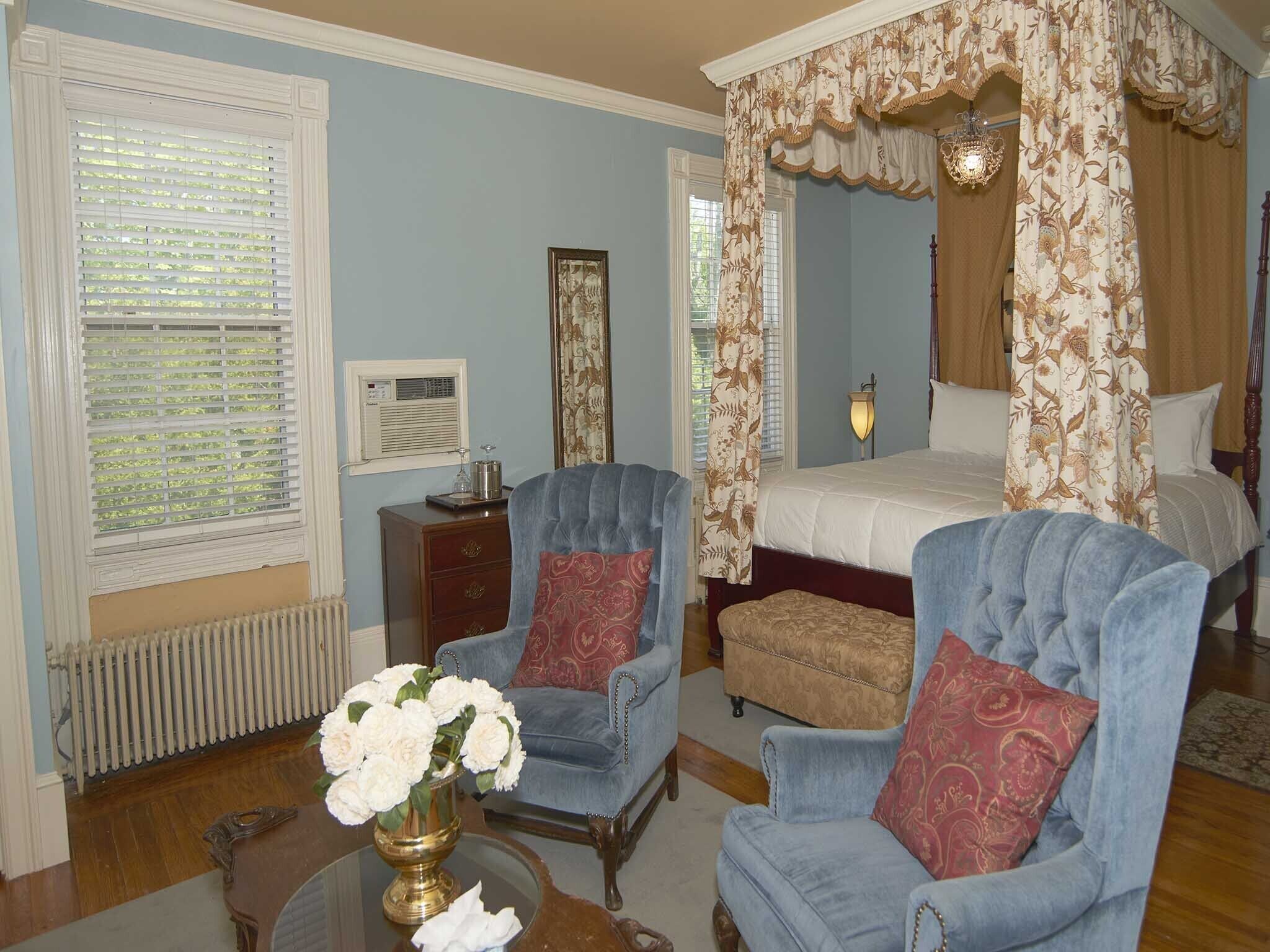 A bedroom with a four-poster bed, two armchairs, a nightstand, a radiator, and a vase of flowers.