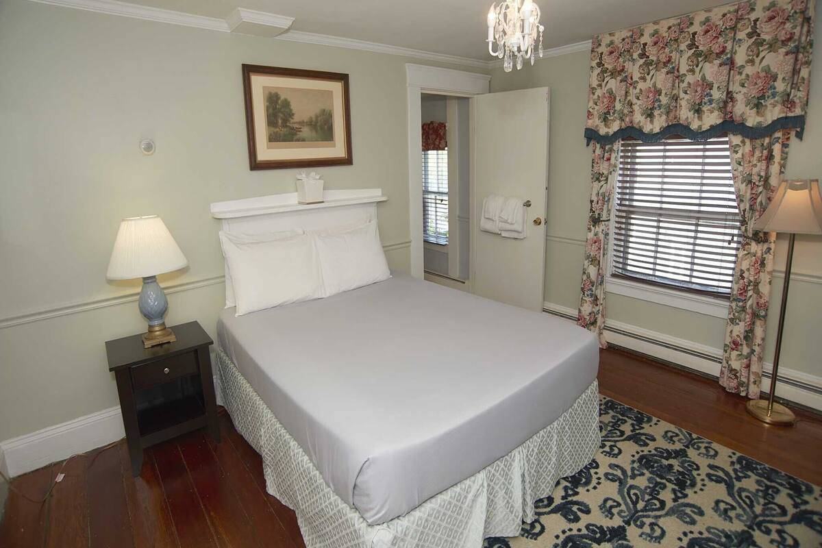 A neatly arranged bedroom with a bed, a nightstand, a lamp, a chandelier, a door, and a window with floral curtains.