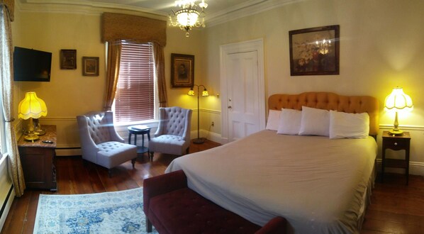 Une chambre d’hôtel avec un grand lit, deux fauteuils, une petite table, une lampe sur pied, un tableau et un lustre.