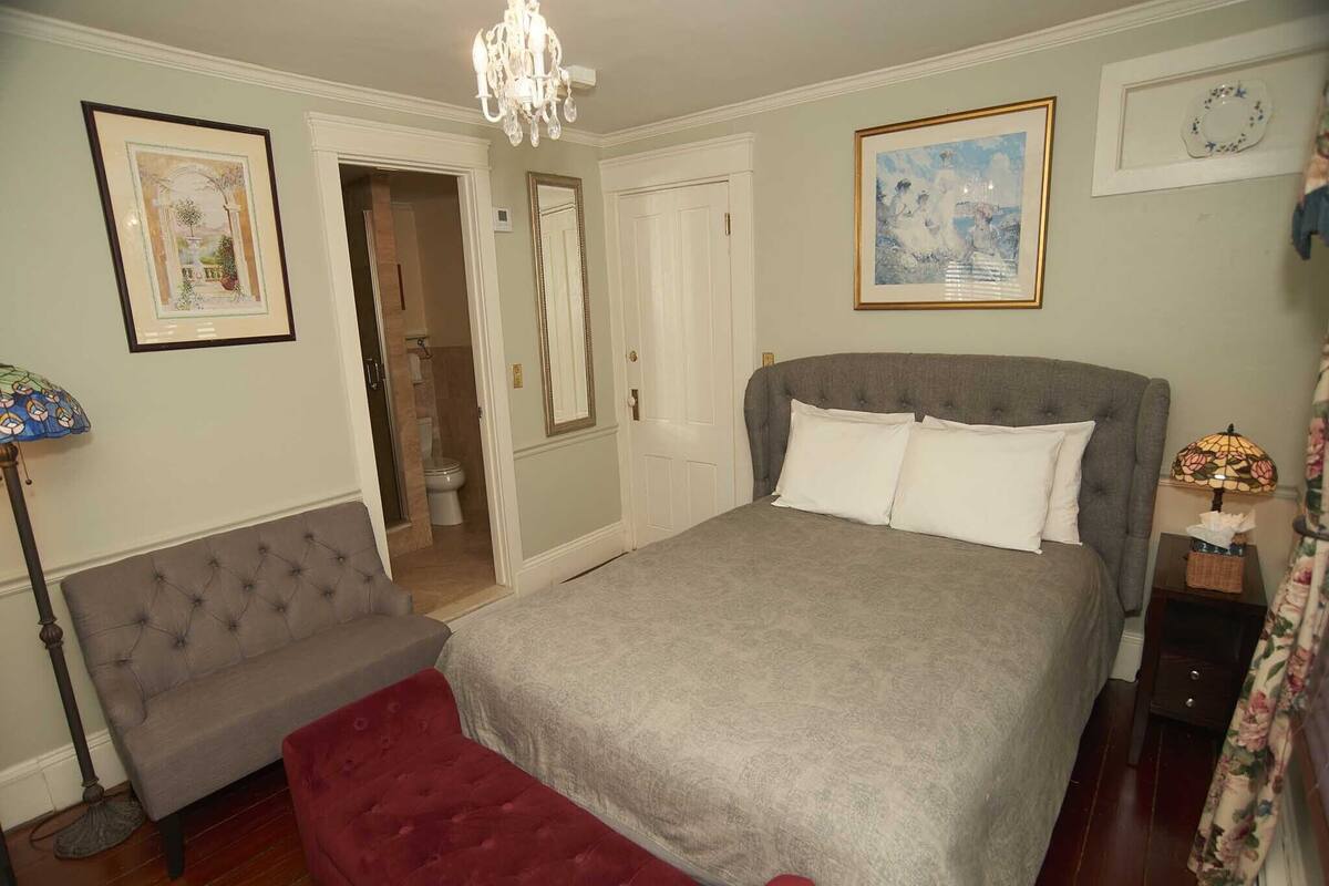 A bedroom with a large bed, a bench, a chandelier, and a door leading to a bathroom.