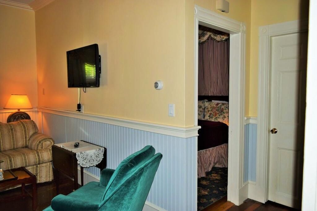 A room with a sofa, a small table with a lace tablecloth, a green chair, a television mounted on the wall, and a door leading to another room.