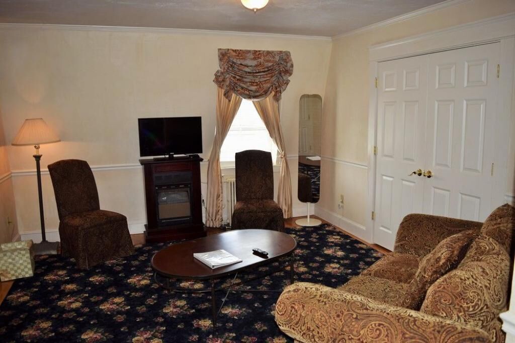 Habitación de hotel con alfombra de motivos florales oscuros, dos sillones, un sofá, un televisor y una puerta grande de color blanco.