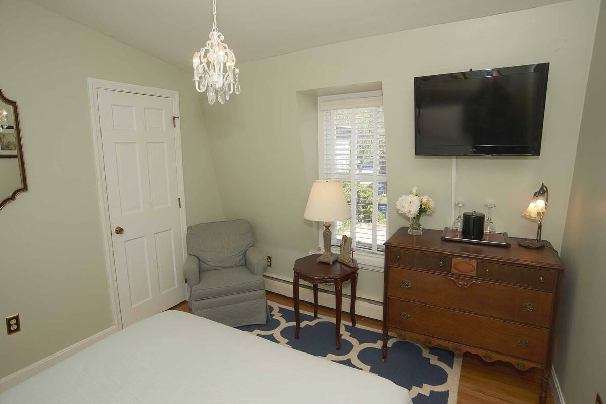 A bedroom with a bed, a dresser, a chair, a lamp, a TV, and a window with blinds.