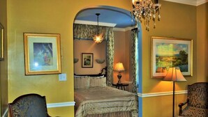 Ein Zimmer mit einem Bett, zwei Stühlen, einer Chandelierleuchtre und gerahmten Bildern an den Wänden.