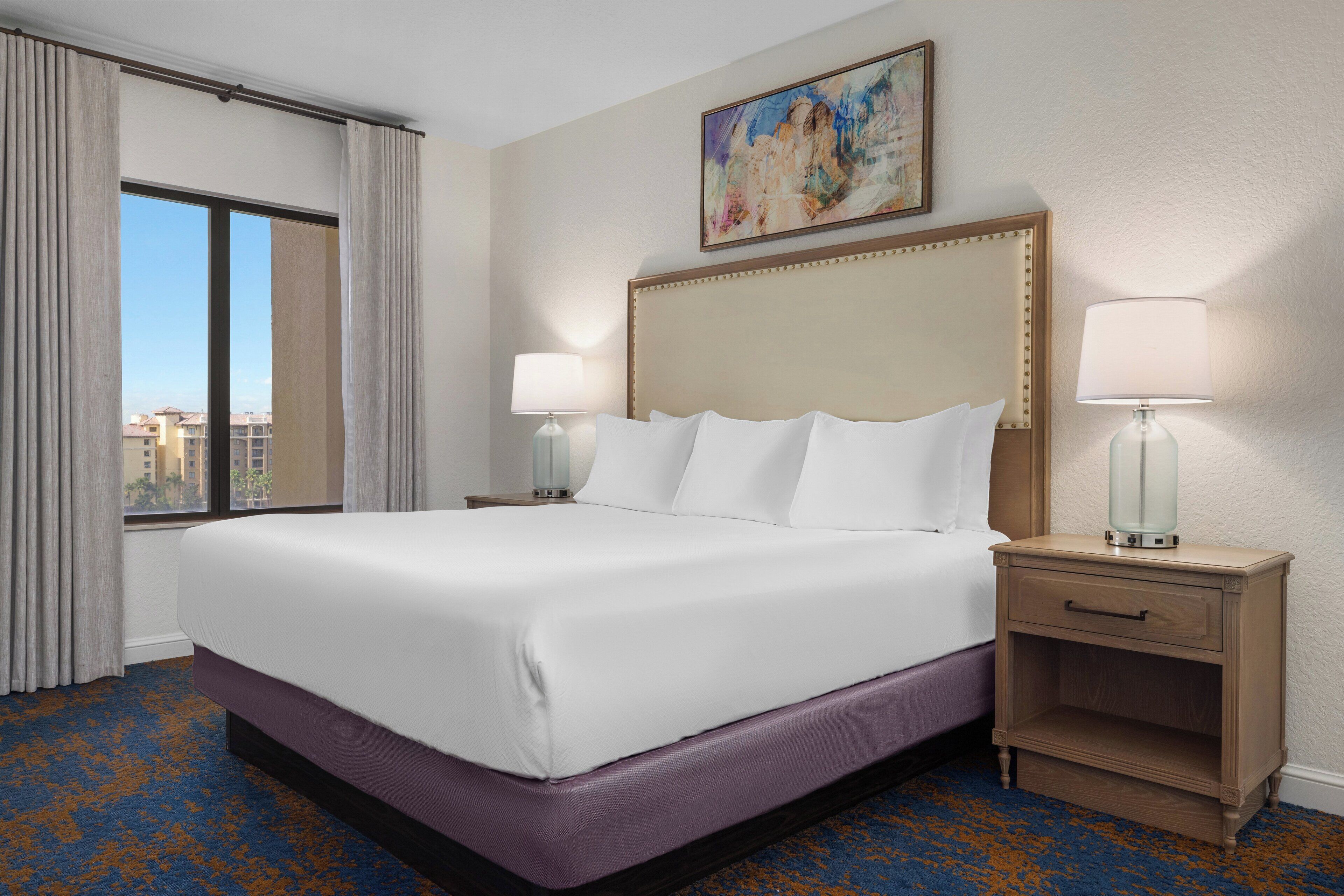 Habitación de hotel con una cama grande, dos mesitas de noche con lámparas, un cuadro en la pared y vistas a la ciudad desde la ventana.