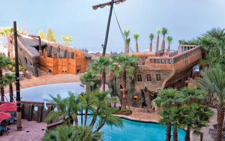 A pool area with a wooden boat structure, palm trees, and a stone building with a balcony.