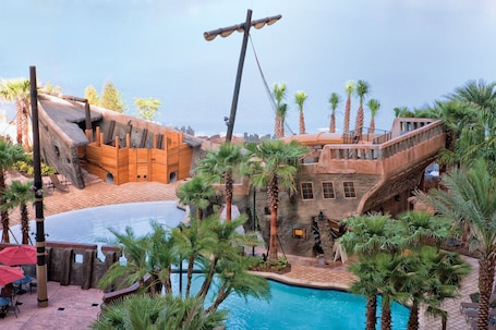A pool area with a wooden boat structure, palm trees, and a stone building with a balcony.