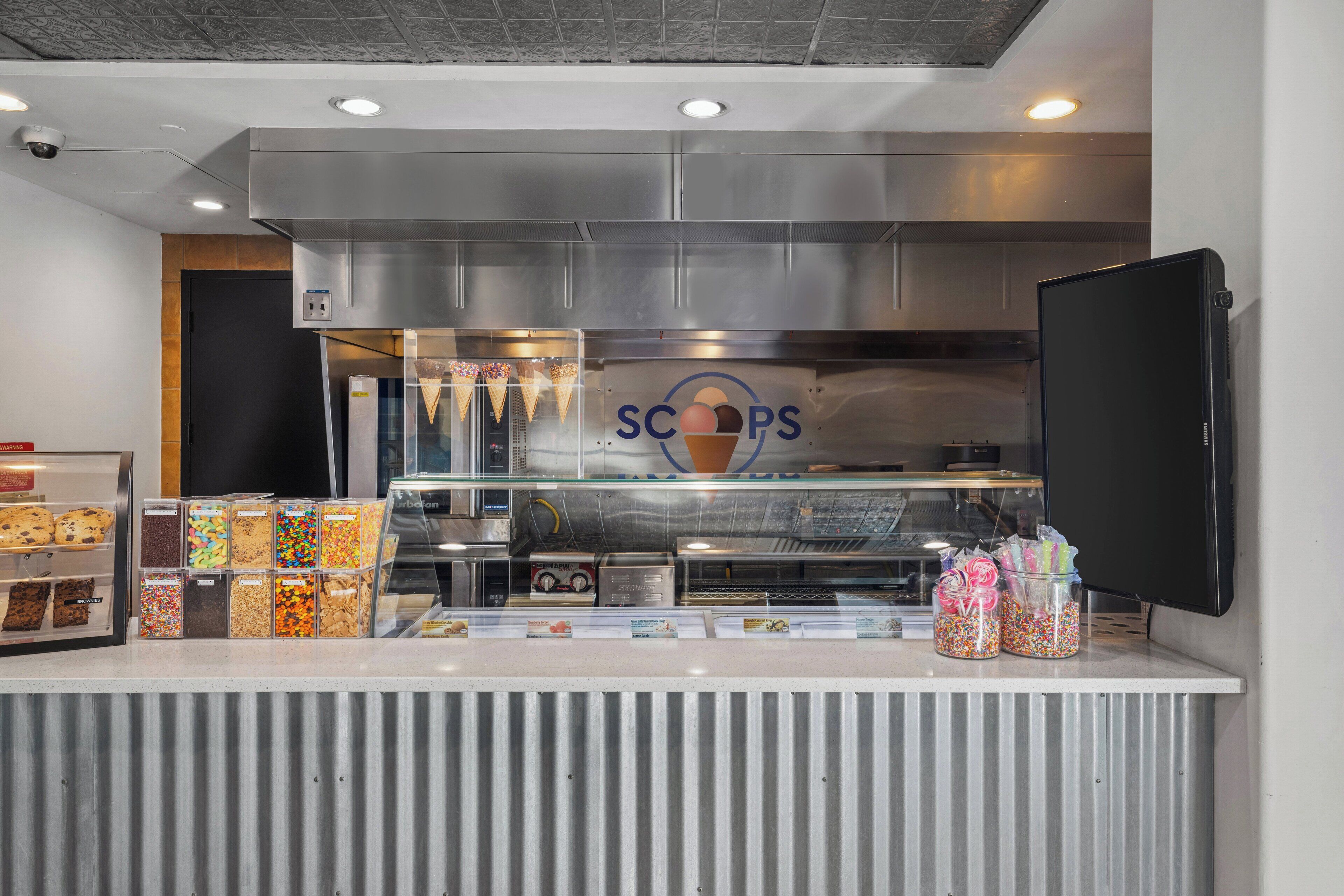 A dessert counter with a variety of ice cream cones and toppings, a freezer, and a sign that reads "SCOPS."