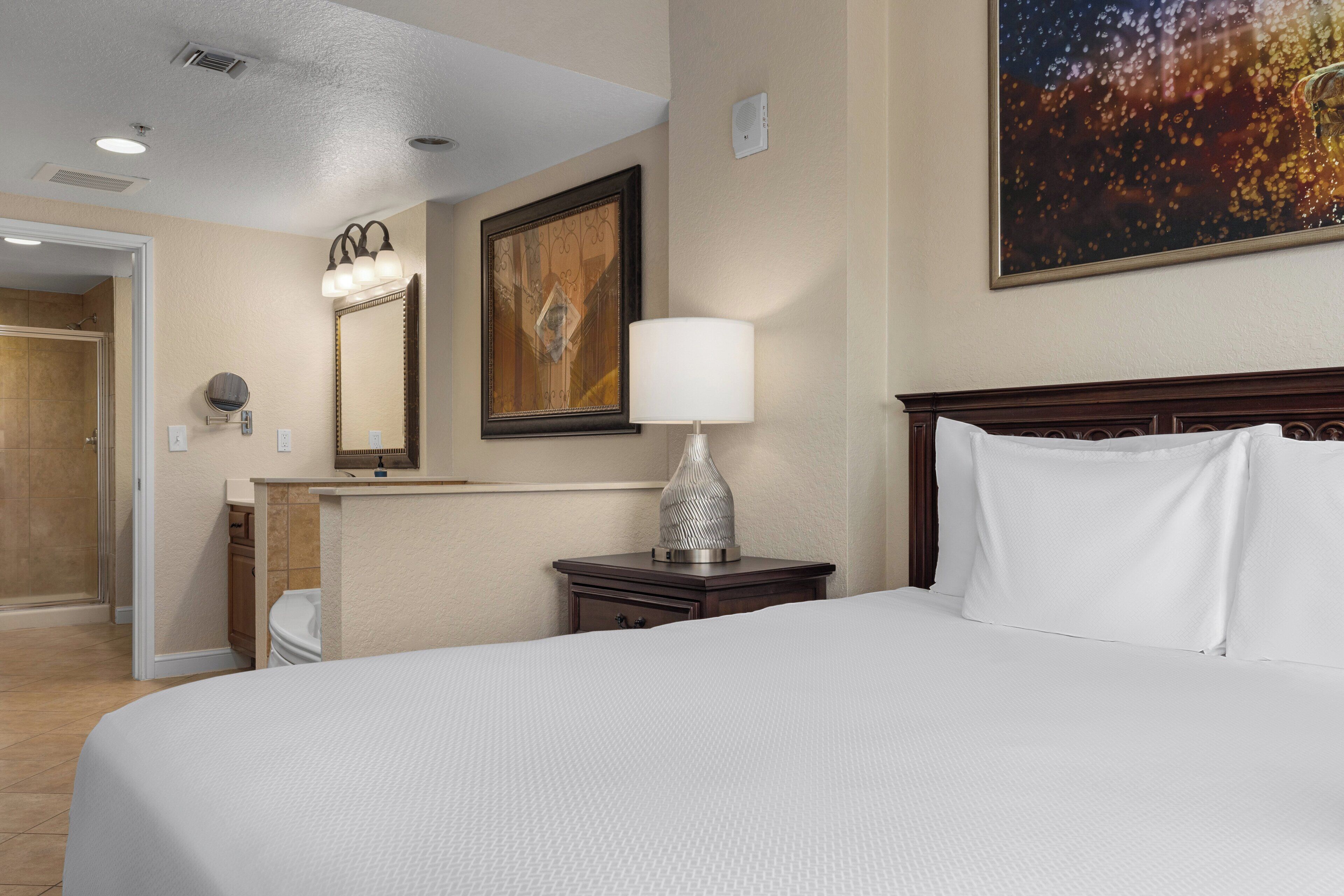 A hotel room with a large bed, a nightstand with a lamp, a mirror, and a painting on the wall.
