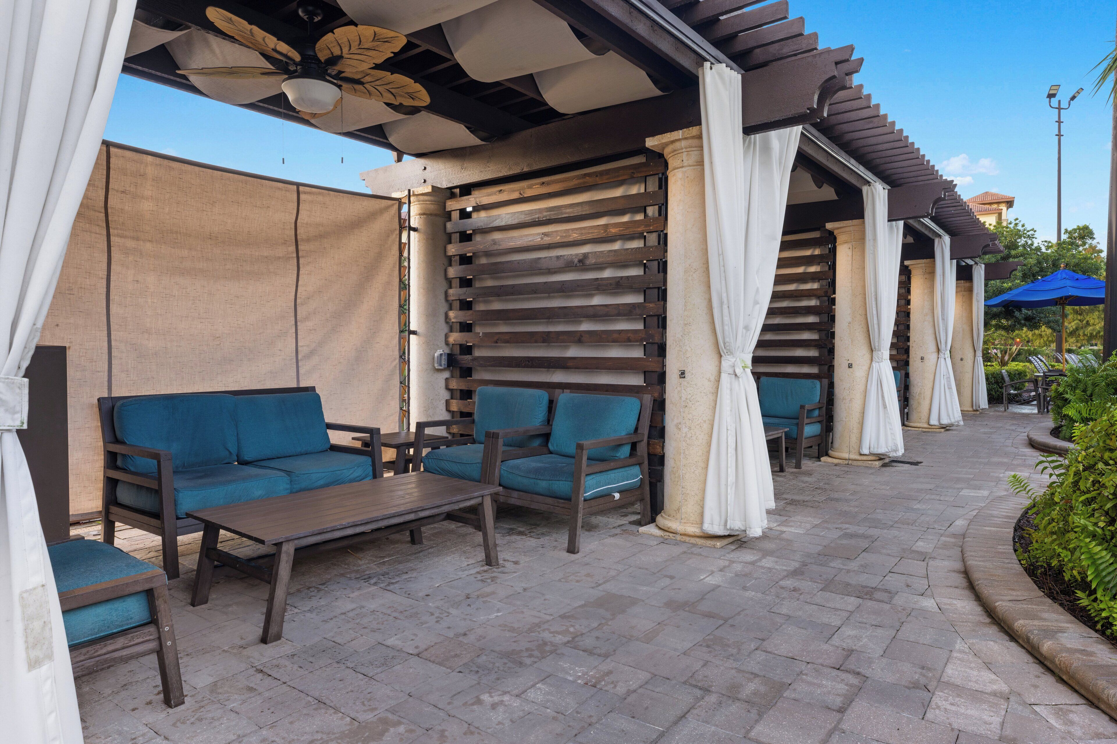 Un patio al aire libre con mobiliario de madera, incluyendo sofás y una mesa, bajo una pérgola con cortinas blancas.