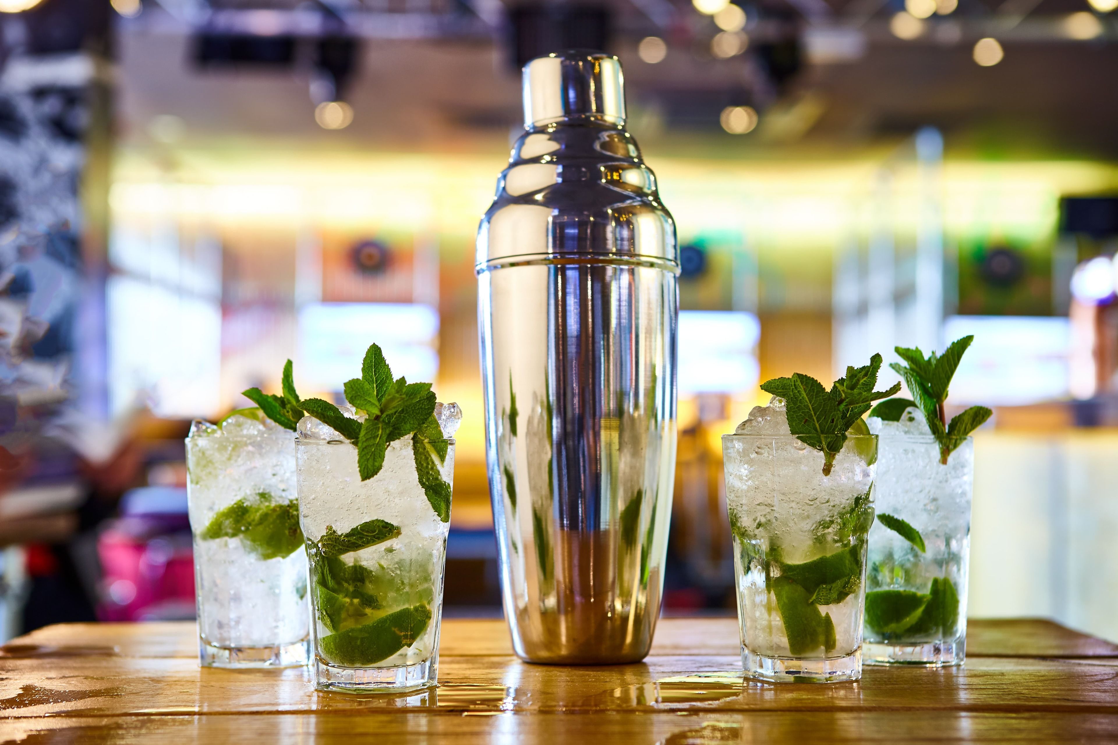 A bar setting with a cocktail shaker and three glasses of a mint-infused beverage.