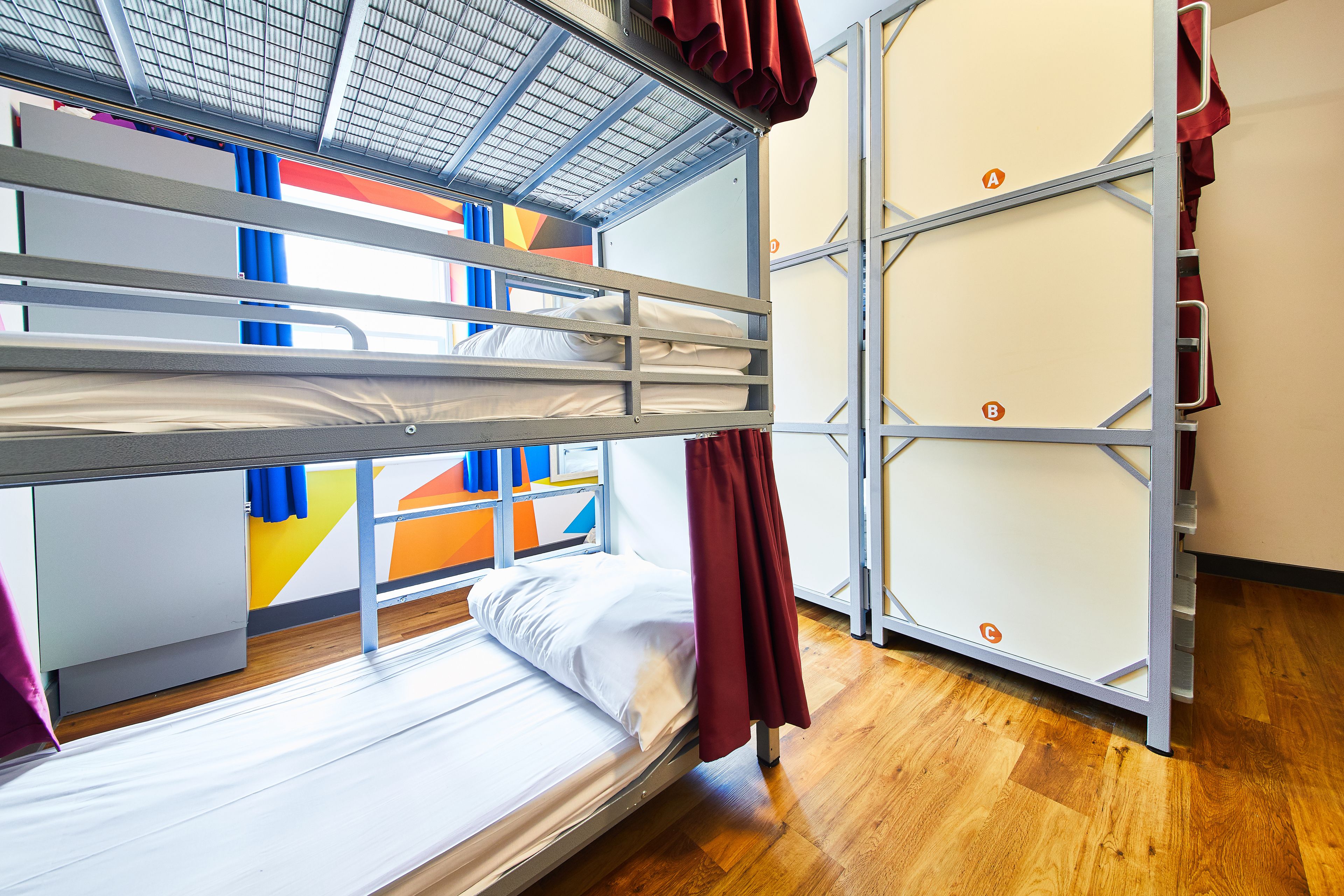 A bunk bed room with a wooden floor, a window with blue curtains, and a colorful wall.