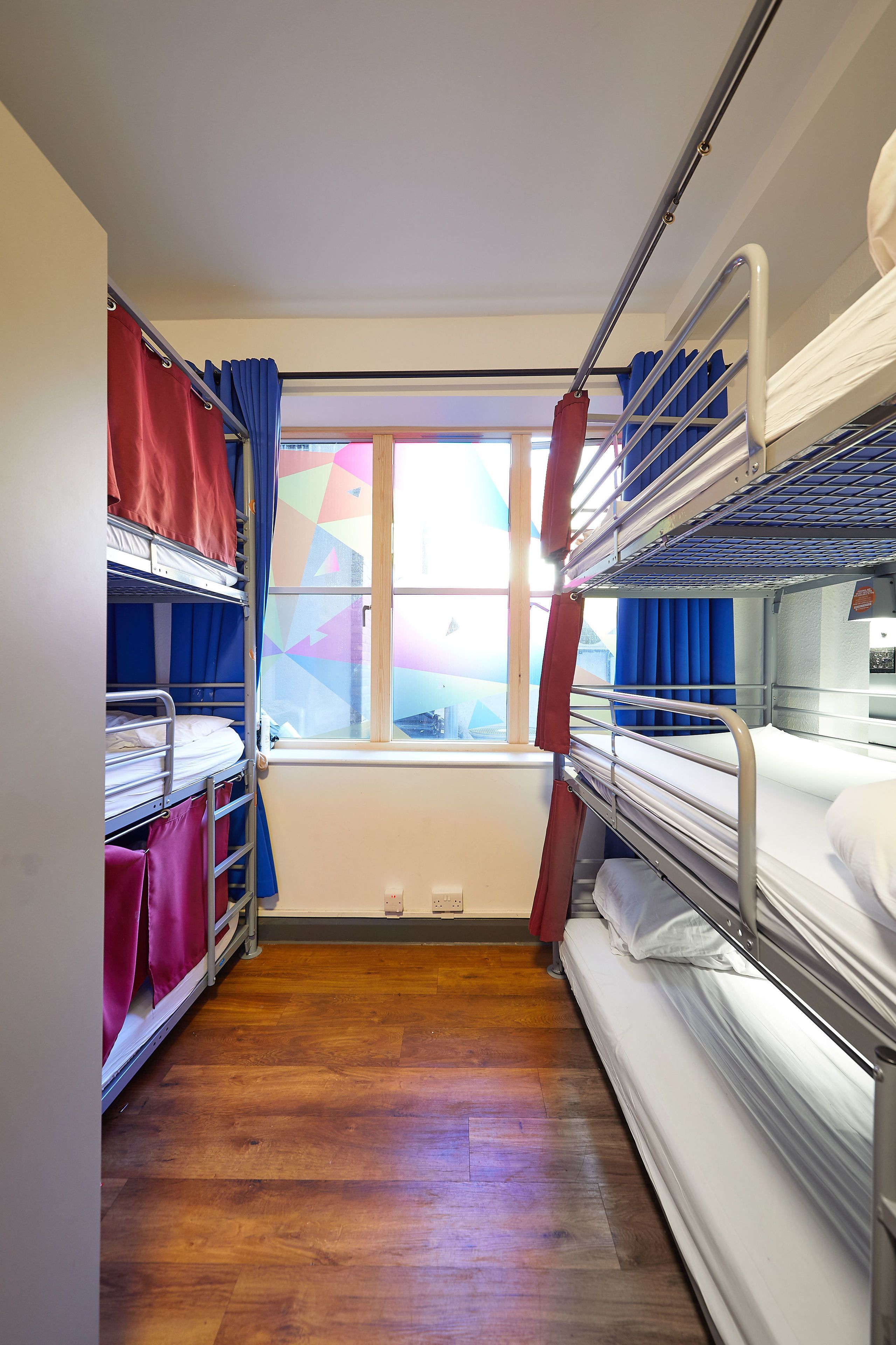 A narrow corridor with bunk beds on either side, a window with curtains, and a wooden floor.