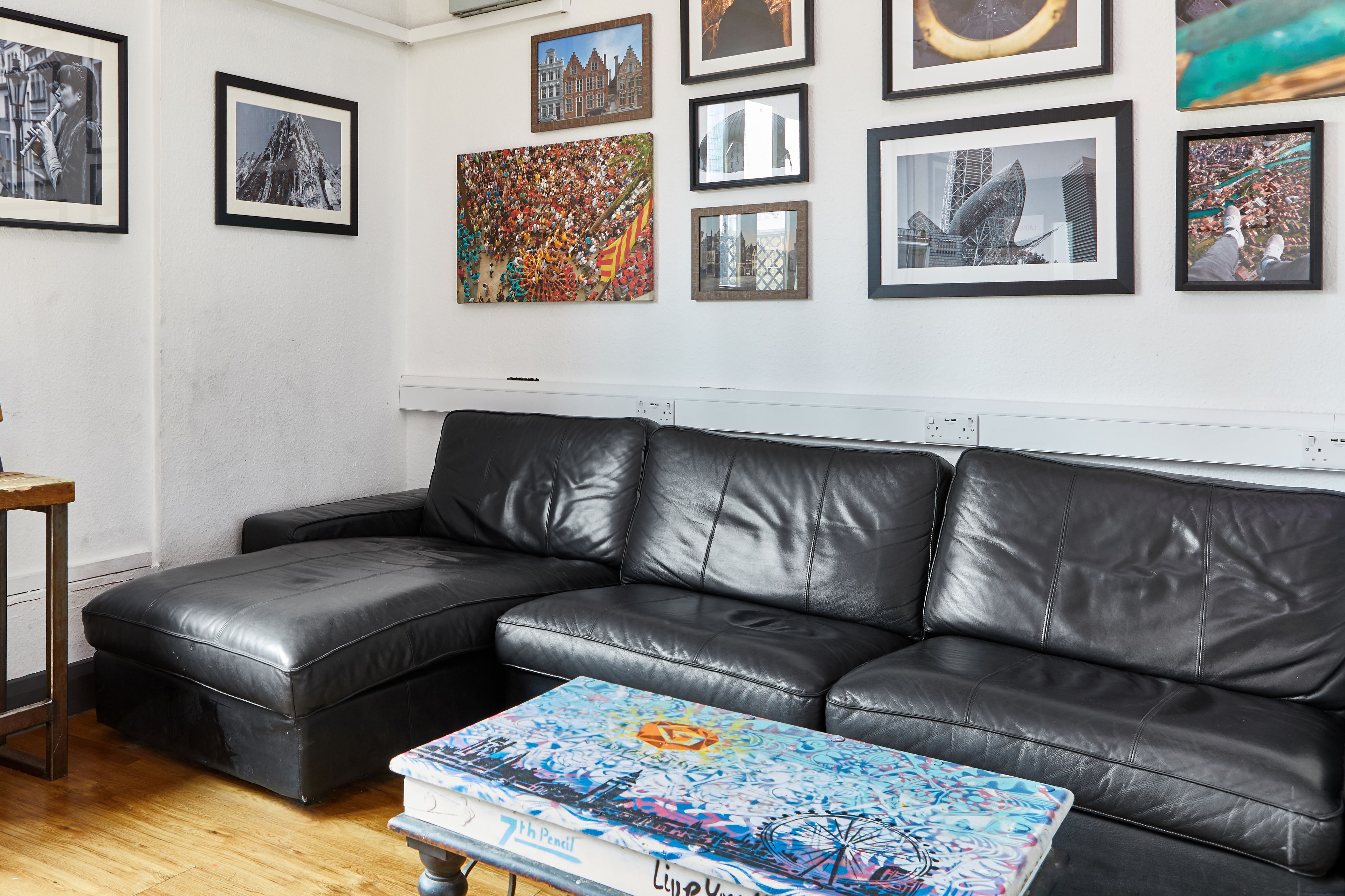 A living room with a black leather sofa, a coffee table with a colorful design, and various framed artworks on the wall.