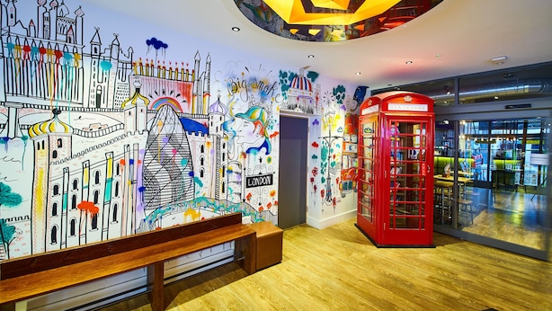 A vibrant mural with cityscapes and iconic landmarks, a red telephone booth, and a wooden bench in a modern interior space. - St Christopher's Village, London Bridge - Hostel (London)