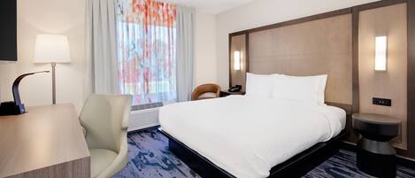 A hotel room with a large bed, a desk, a chair, a desk lamp, a nightstand, and a window with curtains.