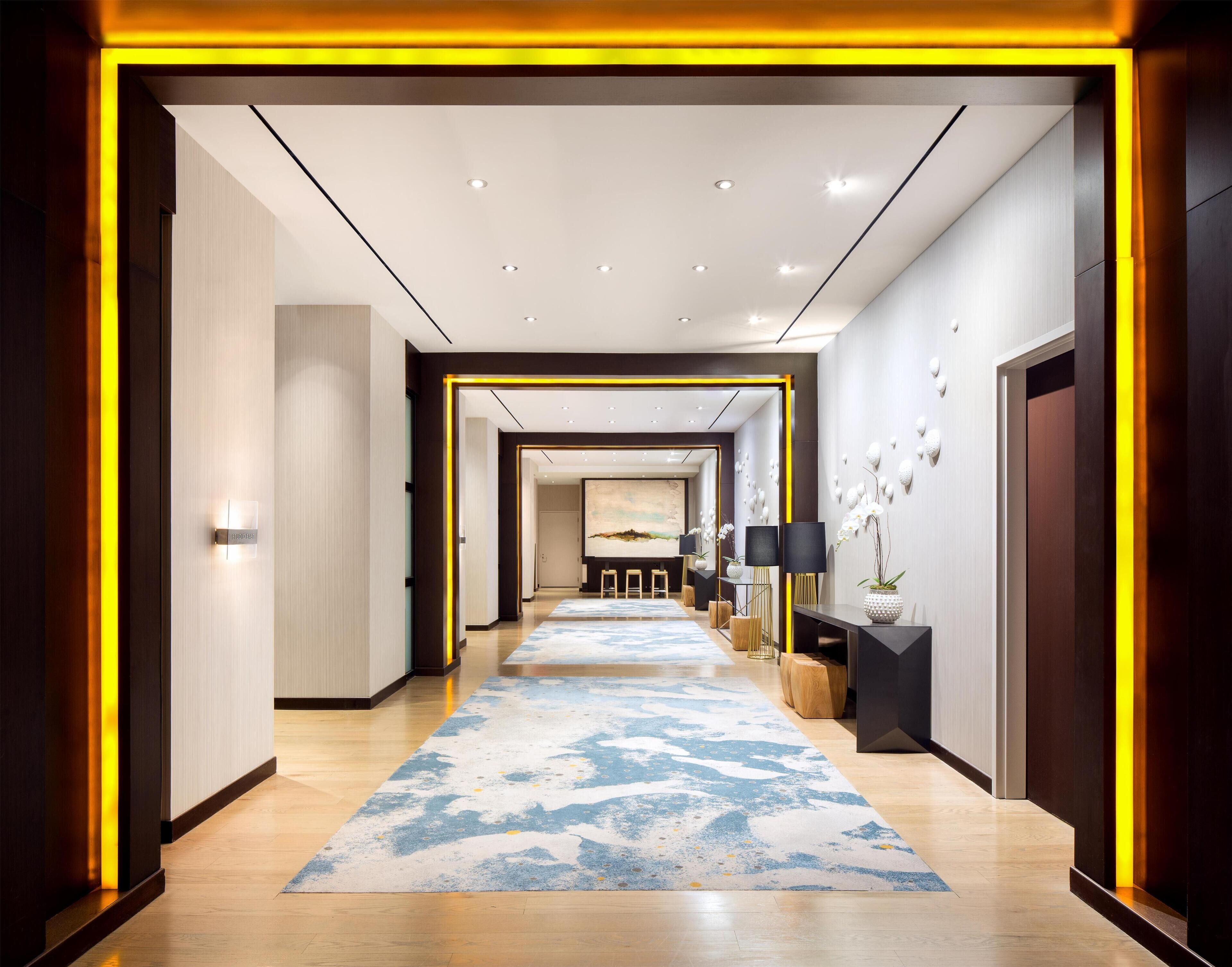 A modern hotel hallway with a light wood floor, a blue and white patterned carpet, and a view into a room with a painting.