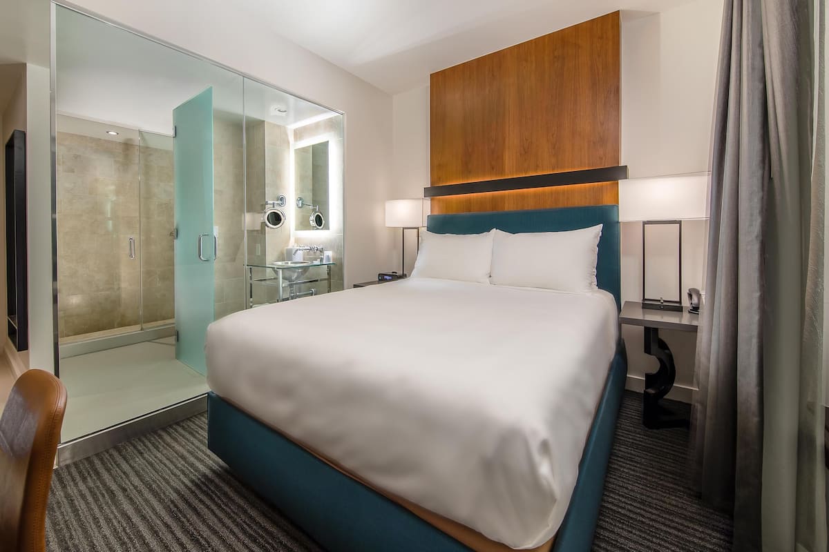 Une chambre d’hôtel comprenant un lit, un bureau, une chaise, une salle de bain avec une douche à parois de verre et une tête de lit en bois.