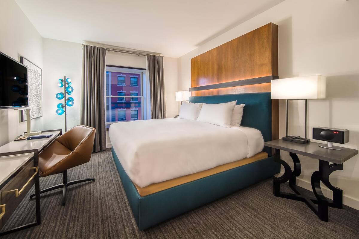 Une chambre d’hôtel avec un lit, un bureau et une chaise, une télévision, une lampe et une vue sur un immeuble par la fenêtre.