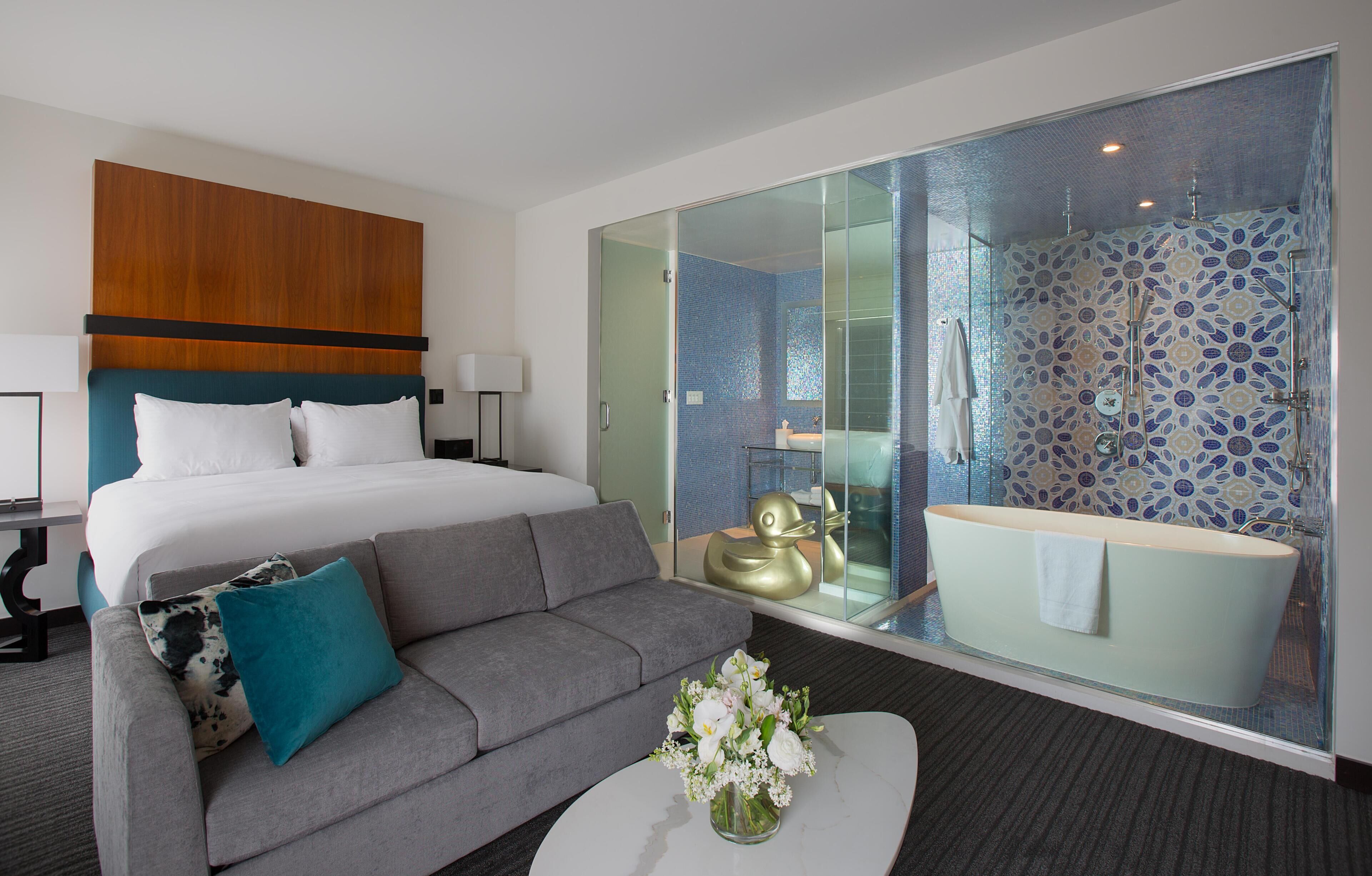 A hotel room with a bed, a sofa, a coffee table with flowers, and a bathroom with a bathtub and shower.