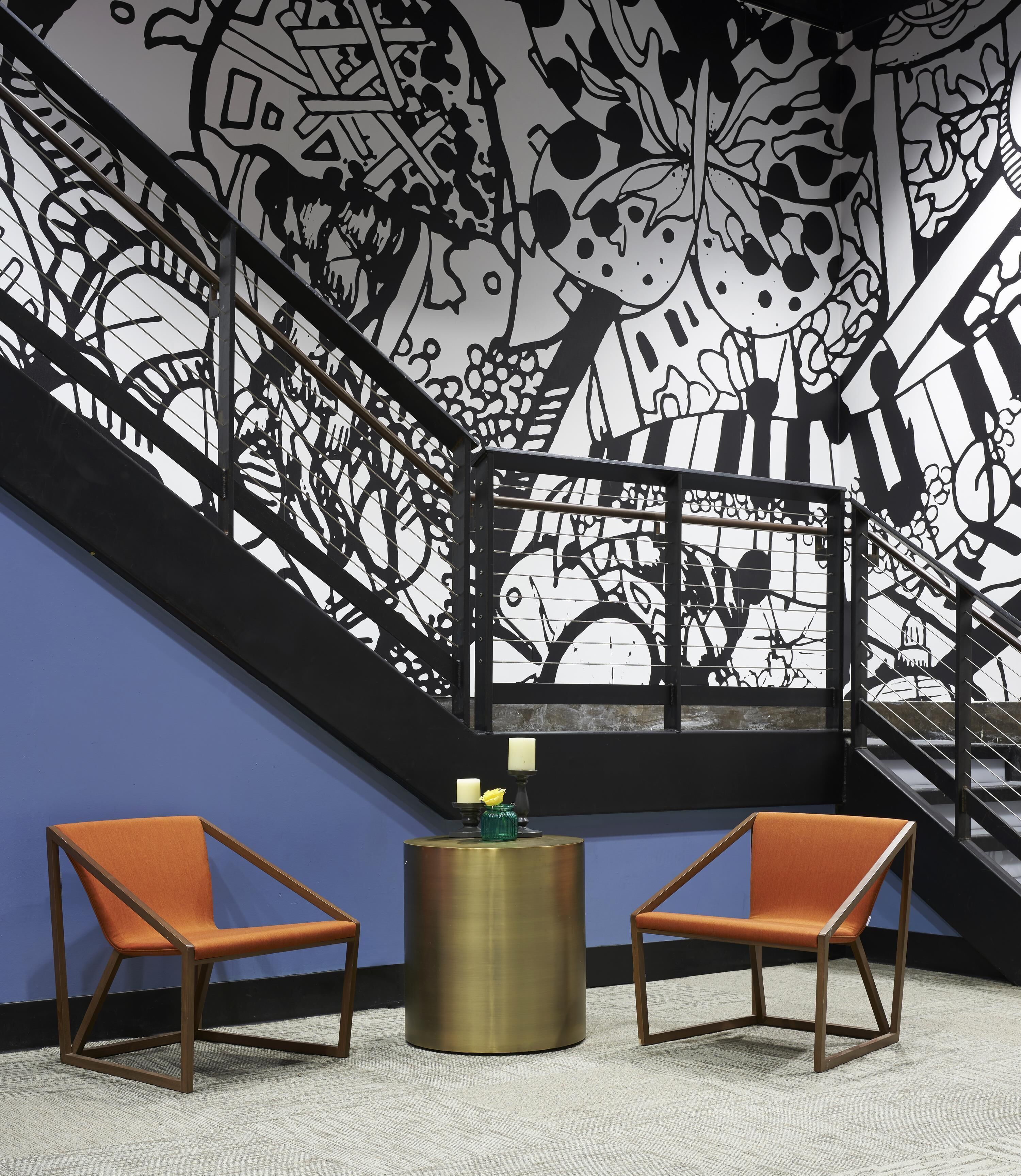 A modern interior with a staircase, two orange chairs, a round table, and a wall with a black and white abstract design.