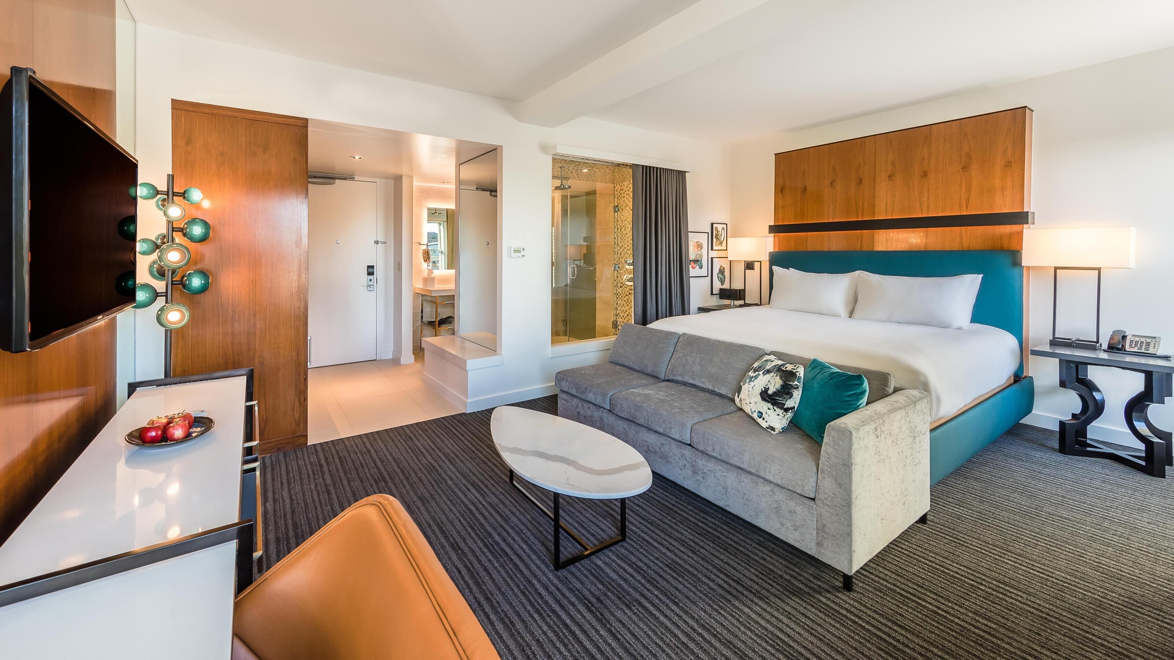 A hotel room with a bed, a sofa, a TV, a desk, and a bathroom visible through a glass door.