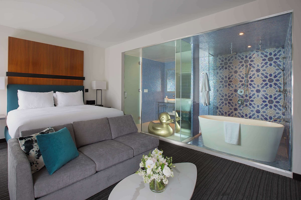 Une chambre d’hôtel moderne avec un grand lit, un canapé gris, une table basse blanche agrémentée de fleurs et une salle de bain fermée par des parois de verre, équipée d’une baignoire et d’une douche.