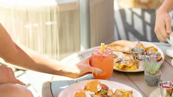 Una mesa de comedor con un plato de huevos benedictinos, cuñas de patata y un vaso de una bebida rosada.