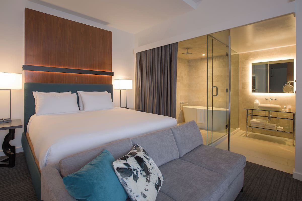 Une chambre d’hôtel moderne avec un grand lit, une douche séparée par des parois en verre, une coiffeuse avec un plateau en verre et un canapé gris agrémenté de coussins décoratifs.