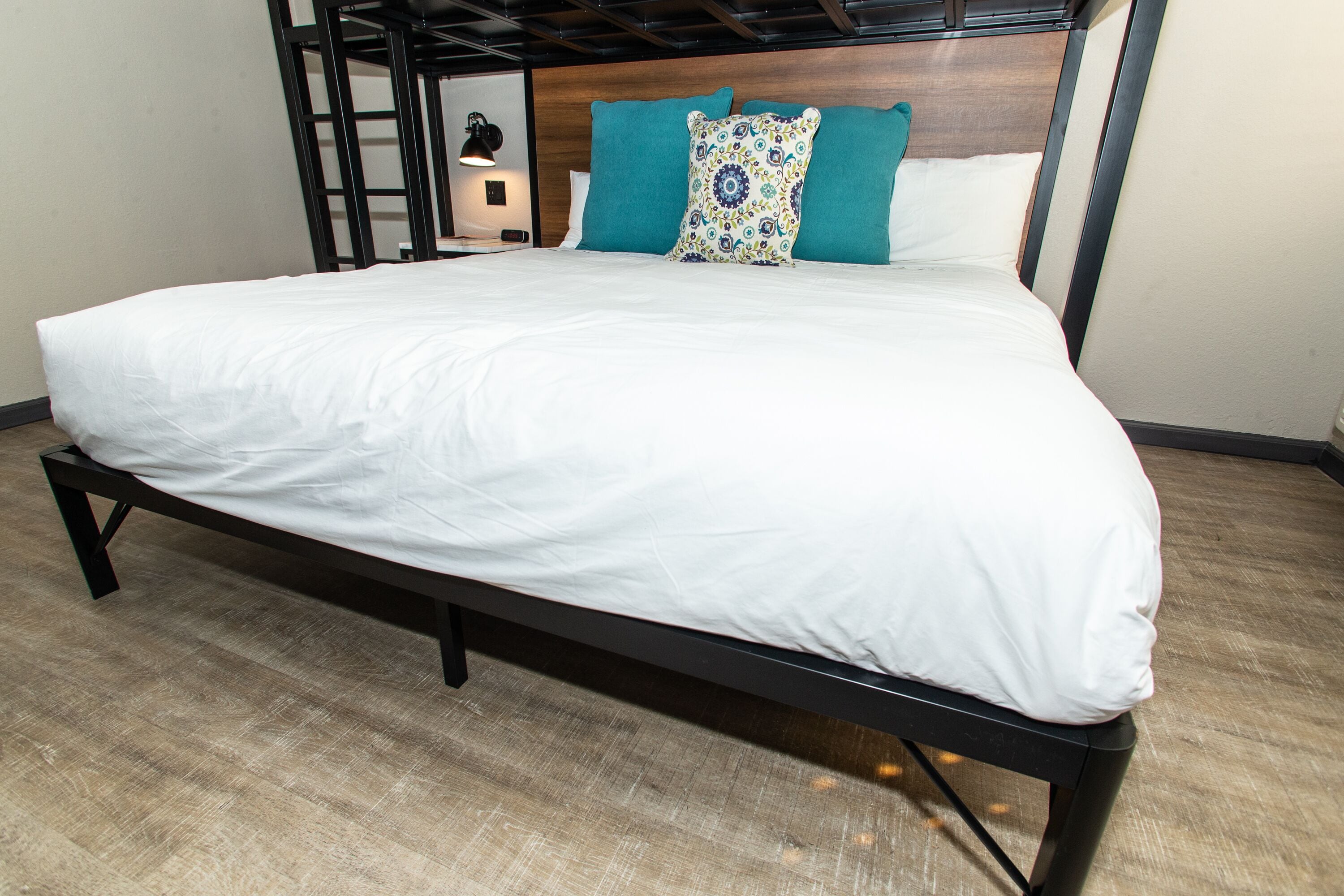 A bed with a white mattress and teal pillows, a wooden headboard, and a nightstand with a lamp.