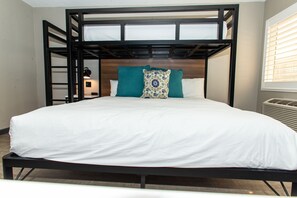 A bunk bed with a white mattress and teal pillow, a bedside table with a lamp, and a window with blinds. - Magnolia Tree Hotel - Near Anaheim Convention Center (Anaheim)