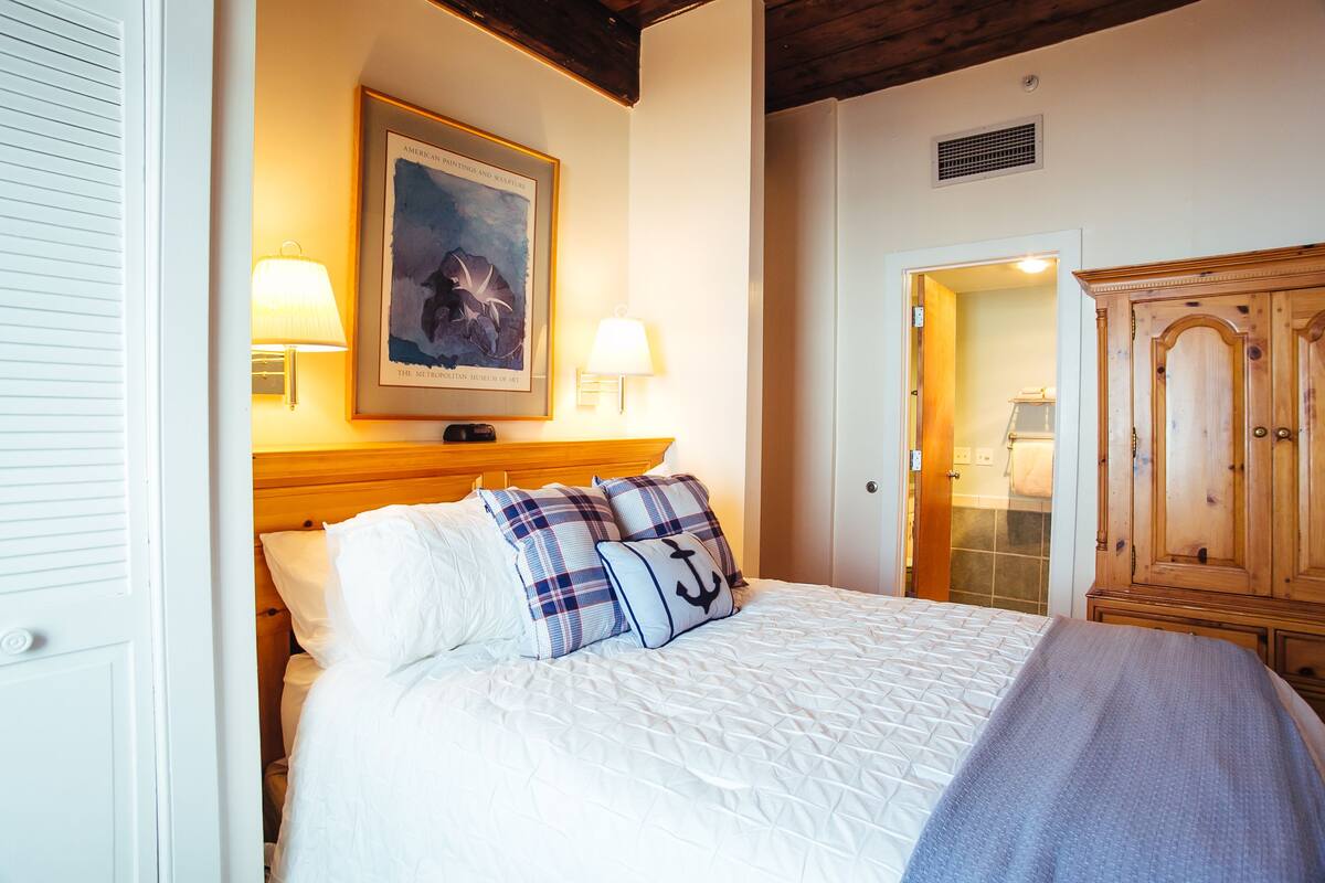 Una cama bien tendida con ropa de cama blanca y almohadas azules y blancas, un cuadro enmarcado sobre la cama y un armario de madera a la derecha.