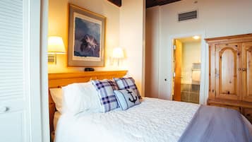 A neatly made bed with white bedding and blue and white pillows, a framed painting above the bed, and a wooden wardrobe to the right.
