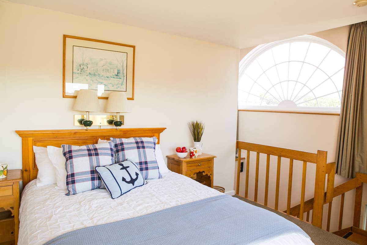 Un dormitorio ordenado con una cama grande, dos lámparas de noche, un cuadro enmarcado en la pared, una mesita con una planta y un florero, y una ventana con cortinas.