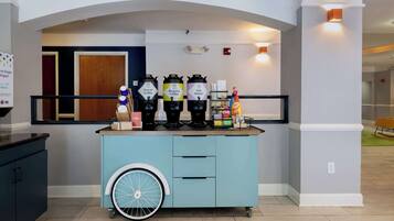 A self-service beverage station with decaf coffee, regular coffee, and hot water options, along with a hand sanitizer dispenser.