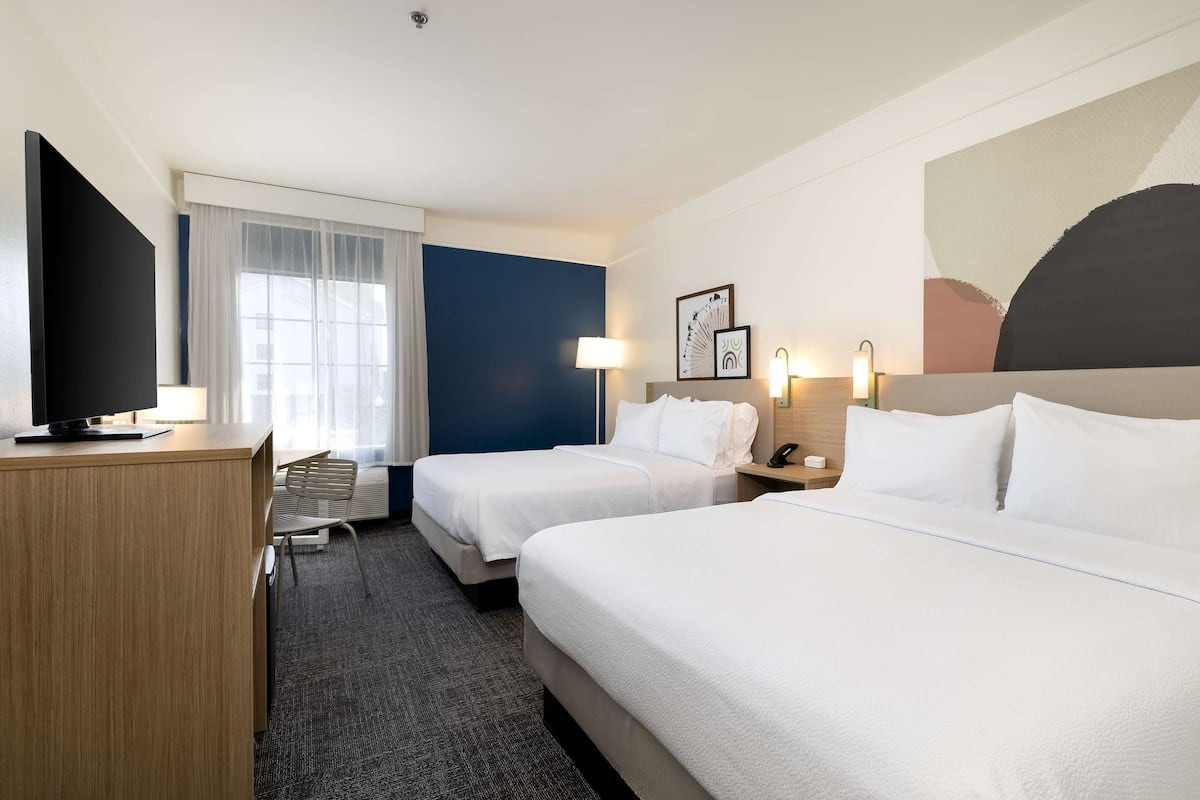 Habitación de hotel con dos camas, un escritorio, un televisor y una ventana con cortinas traslúcidas.