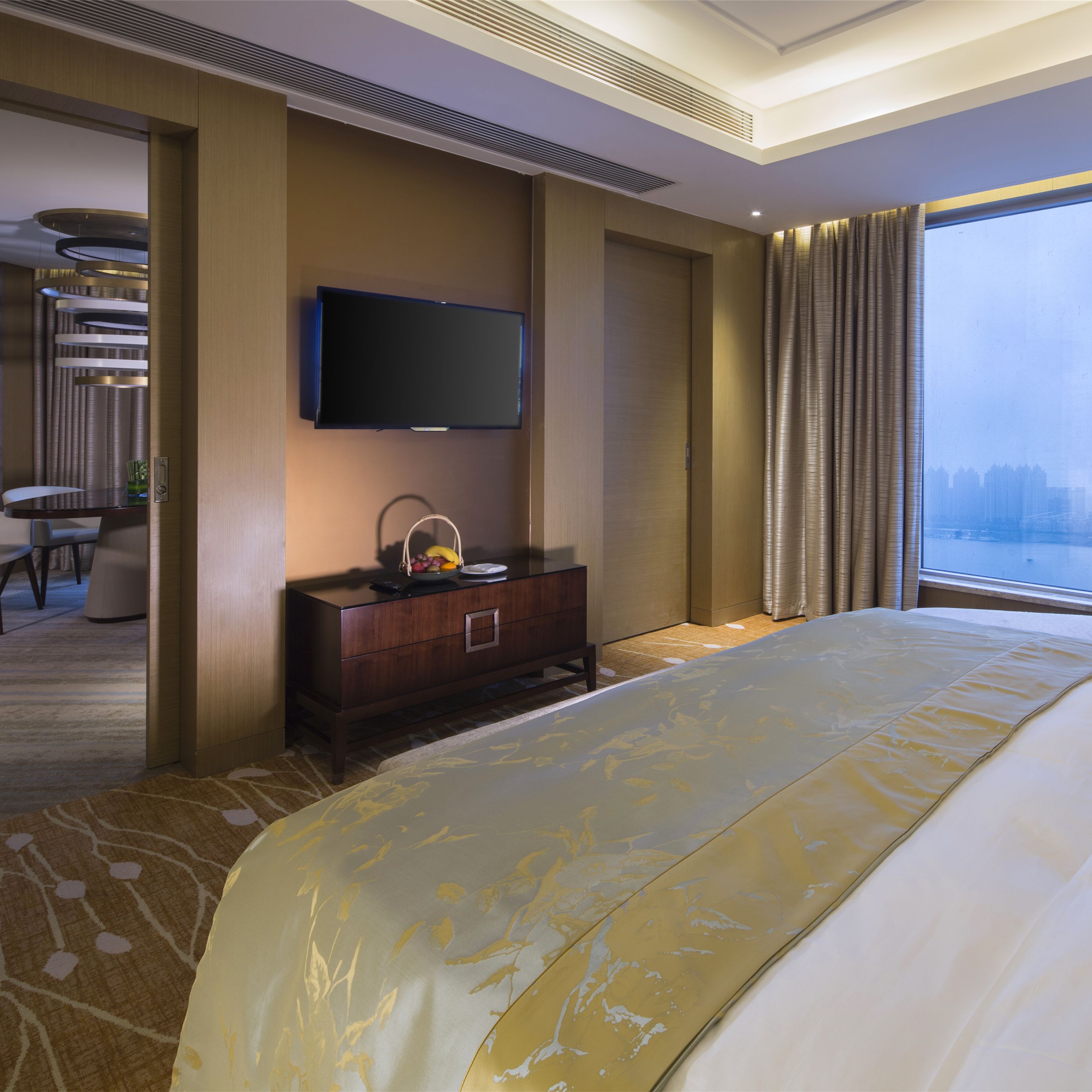 A hotel room with a large bed, a flat-screen TV mounted on the wall, a wooden dresser, and a view of the city through the window.